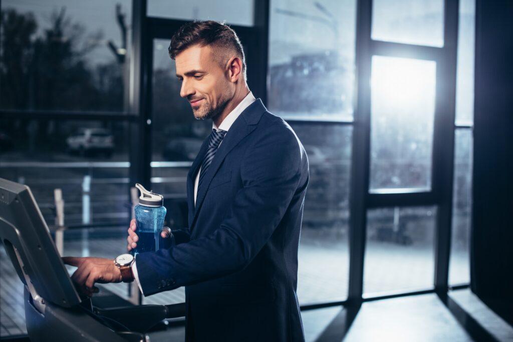 handsome businessman in suit exercising on treadmill and holding sport bottle in gym